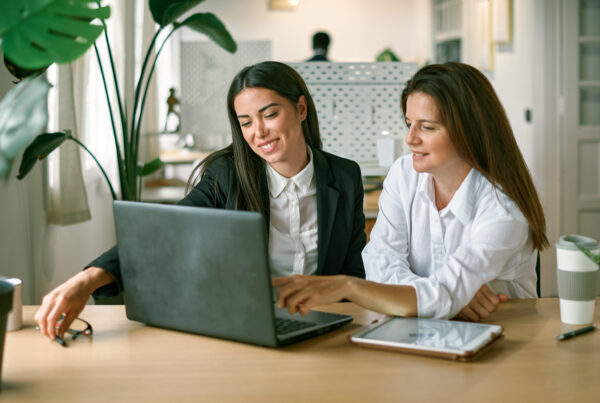 2 jeunes femmes souriantes sont installées à un bureau. Devant elles se trouvent un ordinateur portable et une tablette