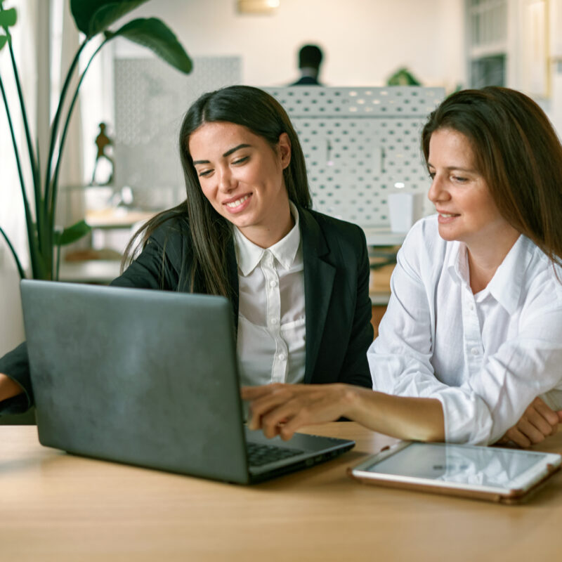 2 jeunes femmes souriantes sont installées à un bureau. Devant elles se trouvent un ordinateur portable et une tablette